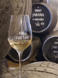 Lustau wine tasting set up, with glasses filled with sherry, and aged barrels in the bodega serving as a backdrop.