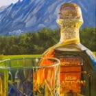 Monica Marquez Gatica's painting depicting a detailed Patron Añejo tequila bottle next to a crystal glass with the Flat Iron mountains in the background.