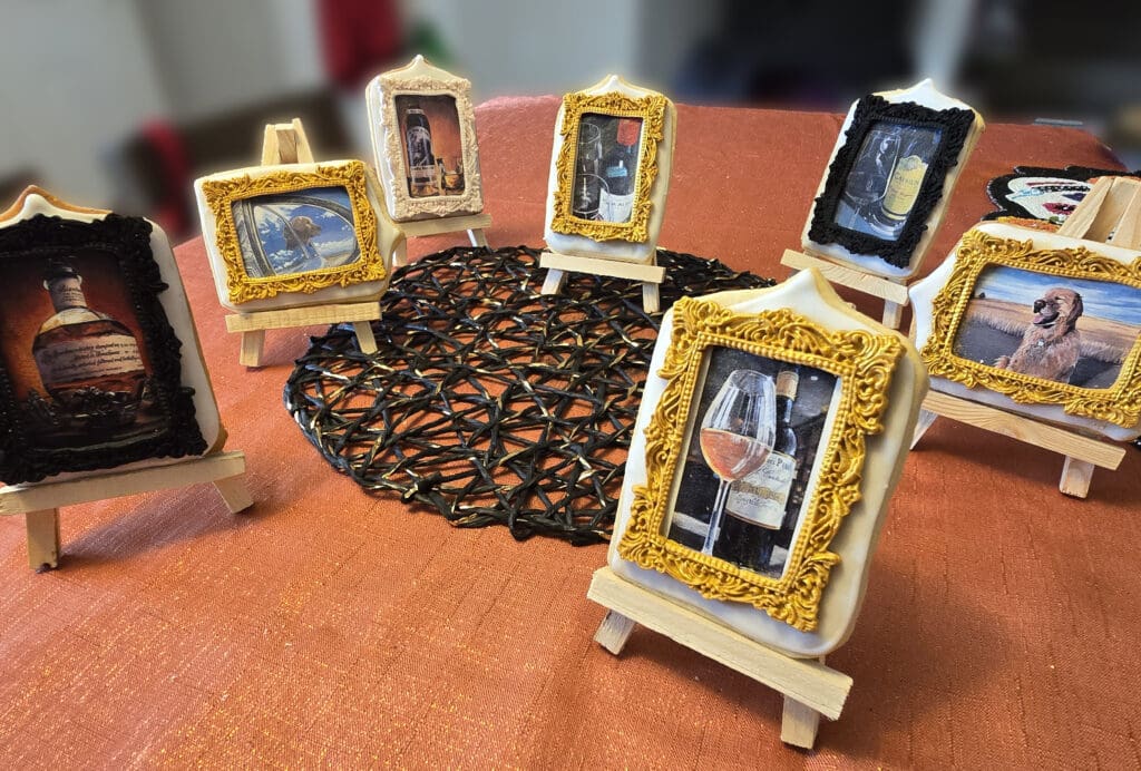 Assorted framed cookies on miniature wooden easels featuring wine bottles, glasses, and playful portraits of a dog.